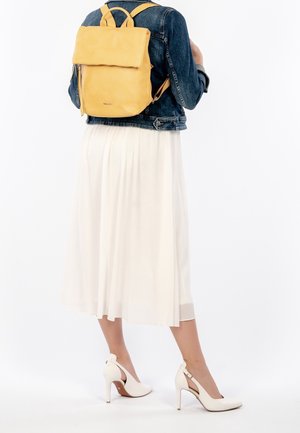 Femme portant une jupe midi blanche, une veste en denim, des talons hauts blancs et un sac à dos en cuir jaune devant un fond blanc.