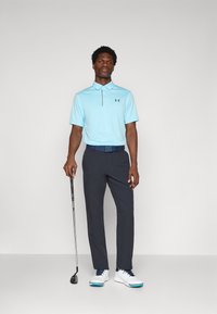 Lichtblauw polo shirt met korte mouwen, gecombineerd met marineblauwe broeken en witte golfschoenen. Een golfclub wordt in één hand vastgehouden.
