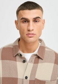 Brown and beige checkered shirt with a textured fabric, featuring a button-up front and a collar. Visible buttons and pocket detail.