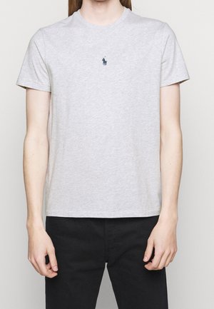 Personne portant un t-shirt à manches courtes gris clair avec un petit logo de joueur de polo sombre sur la poitrine et un pantalon noir.