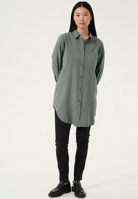 Pika varrukaga, nööpidega särk tuhmunud rohelises kangas esitaskuga; kombineeritud mustade kitsaste pükste ja läikivate mustade kingadega.