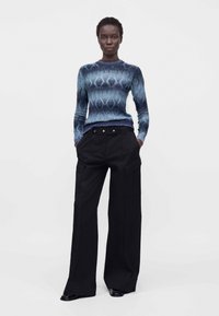 Femme debout, mains dans les poches, portant un haut bleu à manches longues à motifs et un pantalon noir à jambes larges sur fond blanc.