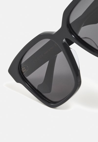 Ochelari de soare supradimensionați, negri, cu o ramă îndrăzneață, geometrică și lentile întunecate. Temelul are inscripția "Gucci" embosată în aur.