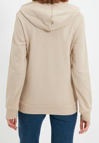 Sudadera beige con capucha, de mangas largas, puños acanalados y bolsillo tipo canguro. Textura suave, corte casual y diseño minimalista.