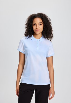 Kobieta o kręconych włosach, ubrana w jasnoniebieską koszulkę polo z krótkim rękawem i czarne spodnie, stojąca na tle jednolitego jasnego tła.