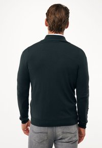 Suéter de manga larga verde oscuro, con cuello y puños acanalados, textura suave, forma ajustada, combinado con jeans grises. Sin patrones ni acentos visibles.