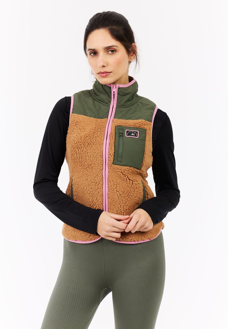 Bruine fleecvest met groene gewatteerde paneel, roze accenten en een ritszak. Draagt over een zwart langemouwen top en geribbelde groene legging.