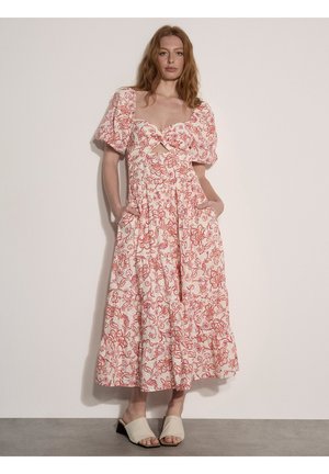 Kobieta ubrana w białą i czerwoną kwiatową sukienkę midi z bufiastymi rękawami i kieszeniami, zestawioną z beżowymi sandałami na słupku z odkrytymi palcami.