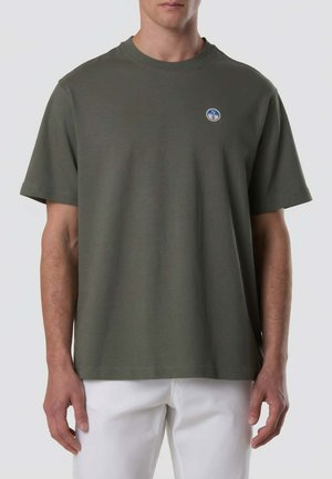 Uomo che indossa una T-shirt a maniche corte verde oliva con un piccolo logo rotondo sul petto, abbinata a pantaloni bianchi su sfondo semplice.