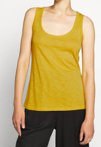 Keltainen tank top, joka on valmistettu kevyestä kankaasta, jossa on syvä kaula-aukko ja leveät hihnat. Topin istuvuus on rento ja materiaali on pehmeä.