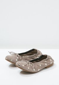 Een paar beige suède ballerina's met zilveren sterrenpatronen en kleine strikjes aan de voorkant, geplaatst op een witte ondergrond.