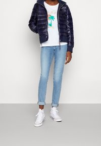 Granatowa kurtka puchowa z poziomym pikowaniem, biała koszulka z nadrukiem z motywem palmy, jasnoniebieskie dżinsy i białe trampki.