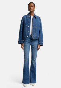 Veste en denim à coupe décontractée, avec de grandes poches avant et des boutons en argent, associée à un jean bleu évasé et des bottines blanches à talons.
