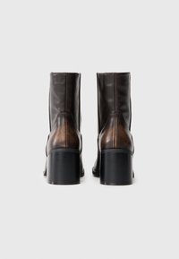 Um par de botins de couro castanho escuro com saltos blocados empilhados, vistos de trás num fundo branco.