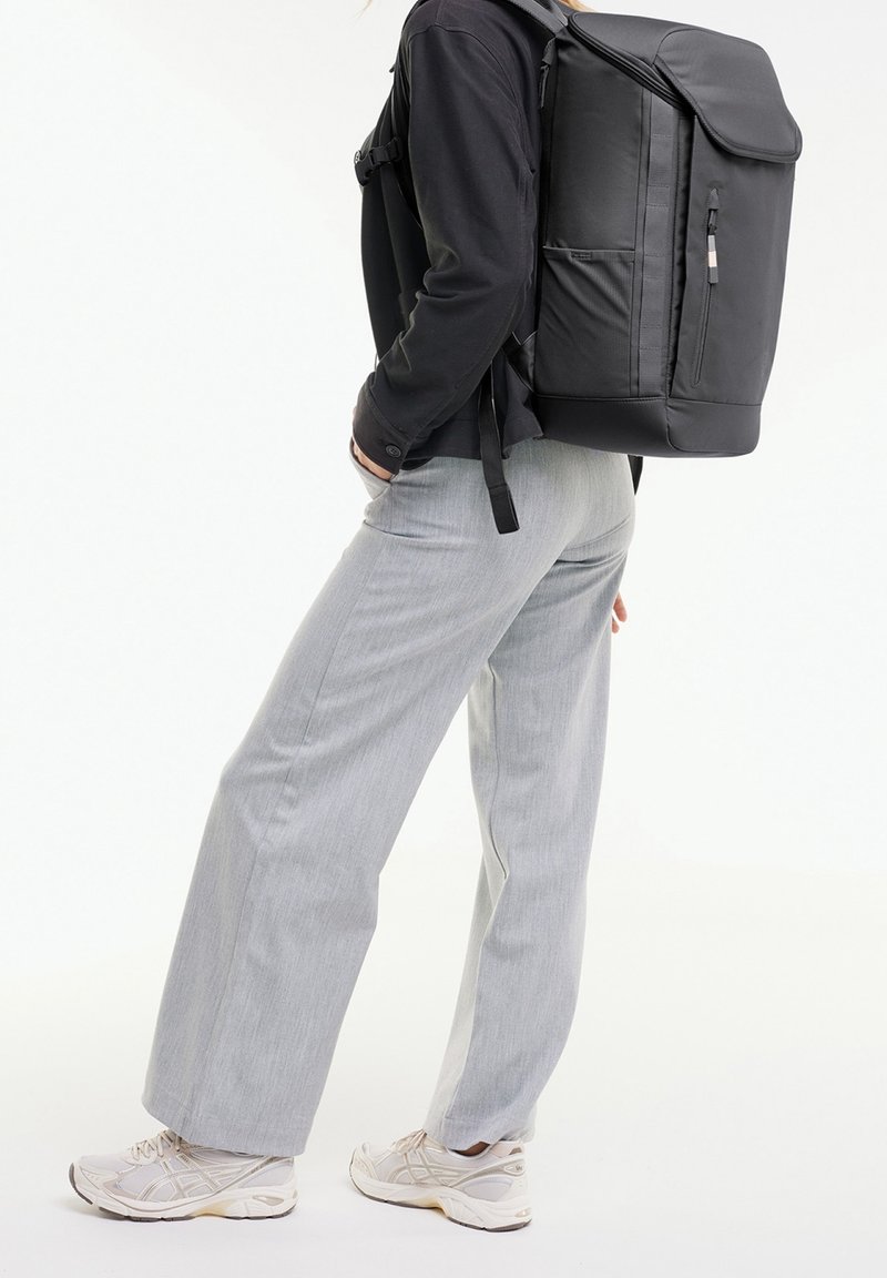 Personne portant un pantalon gris clair, des baskets blanches et un haut noir à manches longues, portant un grand sac à dos noir avec des poches zippées latérales et frontales.