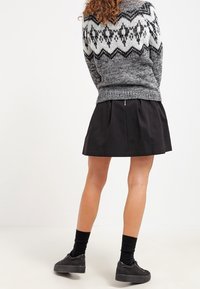 Donna che indossa un maglione grigio con motivo, una gonna plissettata nera, calze nere e scarpe da ginnastica con zeppa nera, in piedi contro uno sfondo bianco.