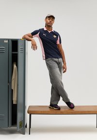 Tamsiai mėlynas ir rožinis polo marškinėliai su logotipu, pilkos džinsinės kelnės ir juodi sportiniai bateliai su violetiniais akcentais. Stovi ant medinės suoliuko prie pilkos spintelės.