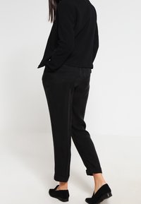 Pantalons noirs sur mesure à texture lisse, ourlets retroussés, assortis à un blazer noir. Des chaussures noires complètent la tenue.