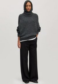 Vrouw in een donkergrijze hoodie, met een witte shirt eronder, zwarte brede broek en zwarte puntige schoenen, staande tegen een effen achtergrond.