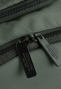 Bolsa de tela texturizada verde que presenta dos cremalleras negras con etiquetas distintivas, asegurada con costuras duraderas.