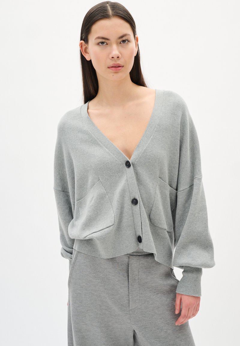 Light grey knit cardigan with a v-neck, front buttons, and two large pockets. The sleeves are slightly oversized and have fitted cuffs.