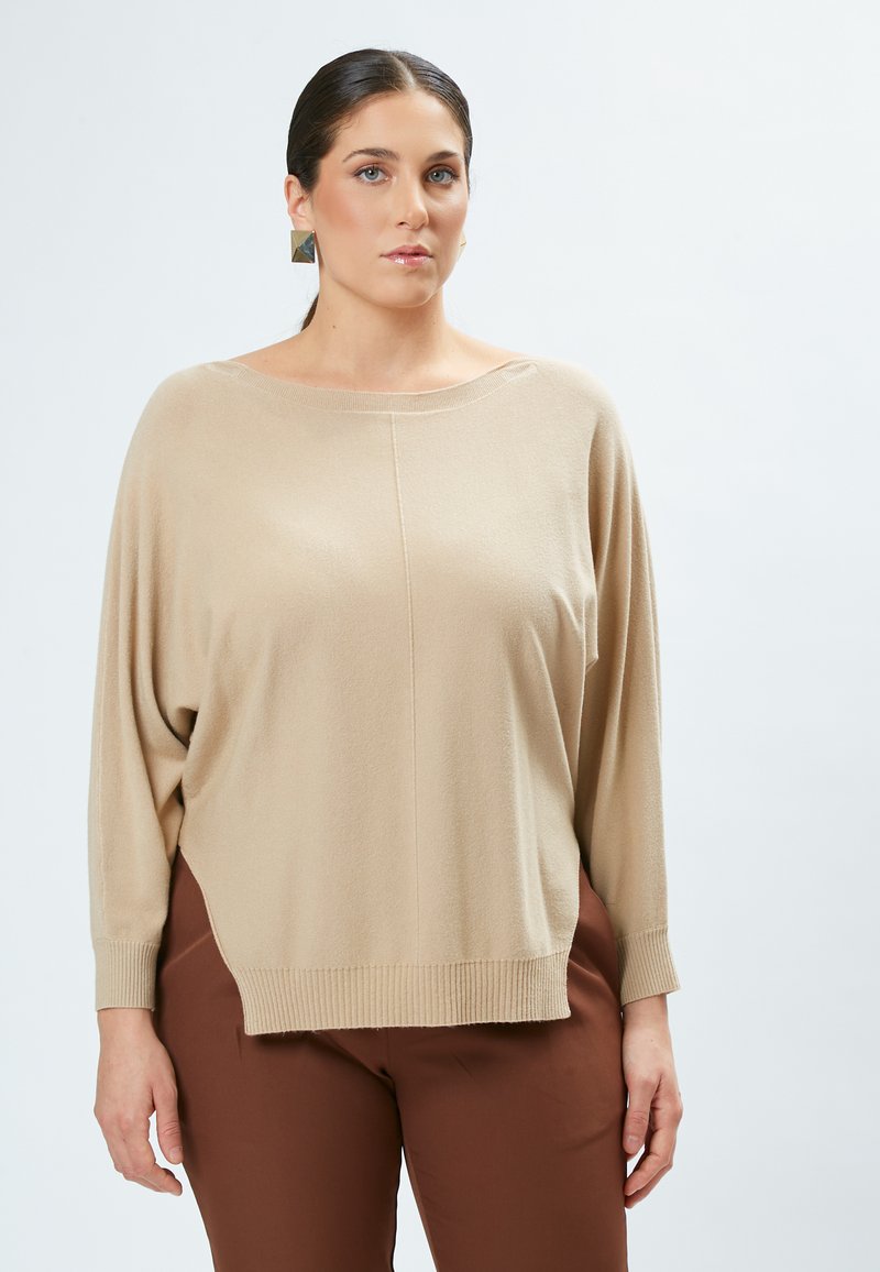 Beige oversized sweater med bådudskæring, ribbede manchetter og en asymmetrisk kant. Glat tekstur og løs pasform, stylet med brune bukser.