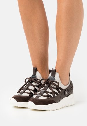 Gambe che indossano sneaker Bally marroni e bianche con suole spesse bianche e lacci ad anello, su sfondo bianco semplice.