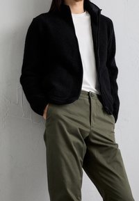 Persona vistiendo una chaqueta negra con cremallera, una camisa blanca y pantalones verde oliva, de pie con las manos en los bolsillos apoyado contra una pared gris texturizada.