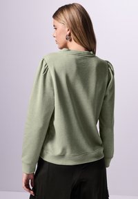 Grön sweatshirt med puffärmar, rund halsringning och ribbade ärmslut. Slät textur och enhetlig färg, stylad med mörka byxor.