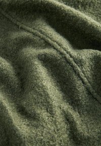 Groene textuurstof met een fuzzy oppervlak, met een zacht, chunky ontwerp en zichtbare stiksels, wat een warme, gelaagde uitstraling creëert.