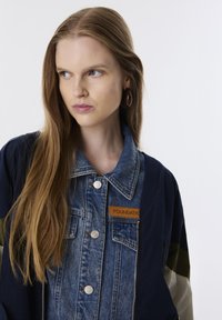 Denim jacket met marineblauwe en olijfgroene accenten, voorzien van een knoopsluiting aan de voorkant, een grote kraag en een stoffen patch met het label "FOUNDATION."