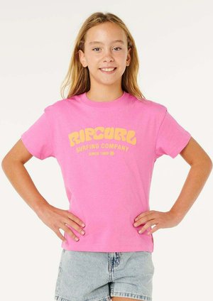 Lachend meisje met lang haar draagt een felroze Rip Curl T-shirt en lichte spijkershorts, staat met handen in de zij tegen een effen achtergrond.