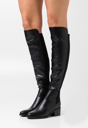 Stivali neri in pelle al ginocchio con tacchi bassi a blocco, indossati su gambe nude, mostrati da davanti su sfondo bianco.