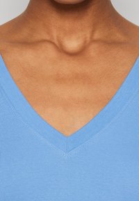 Světlomodrý top s výstřihem do V vyrobený z žebrované tkaniny, který má plochý límec a hladkou texturu. Podrobnosti šití jsou viditelné podél okrajů.