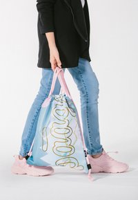 Jasnoniebieski worek na sznurku z złotym abstrakcyjnym wzorem, różowymi akcentami, trzymany przez osobę w czarnej kurtce i jasnoniebieskich dżinsach, ubraną w różowe buty.