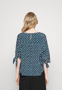 Blouse bleue à motifs avec des manches courtes, dotée de liens aux poignets, d'un col rond et d'une découpe dans le dos. Texture de tissu douce.