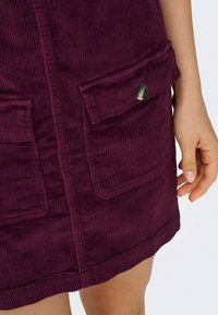 Gros plan d'une jupe en velours côtelé violet avec poches à rabat et un bouton, portée par une personne avec une main visible sur le côté.