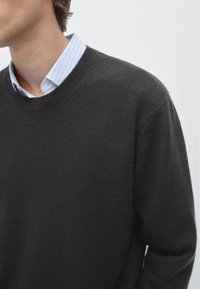 Ciemnoszary sweter z dzianiny z okrągłym dekoltem, noszony na jasnoniebieskiej koszuli w paski. Gładka struktura i luźny krój, z prążkowanymi mankietami.