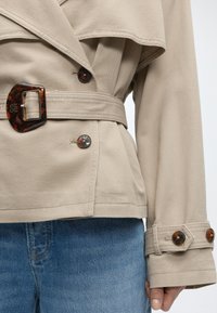 Μπεζ μπουφάν με ζώνη και κουμπιά από χελωνόκερο πάνω από μπλε τζιν, που δείχνει το κομμένο κορμό και το δεξί χέρι με γυμνό βερνίκι νυχιών.