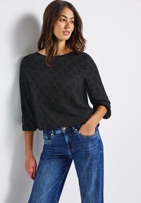 Zwarte blouse met een geometrisch bruin patroon, boot-halslijn en driekwartmouwen, gedragen met blauwe denim jeans. Textuurstof.