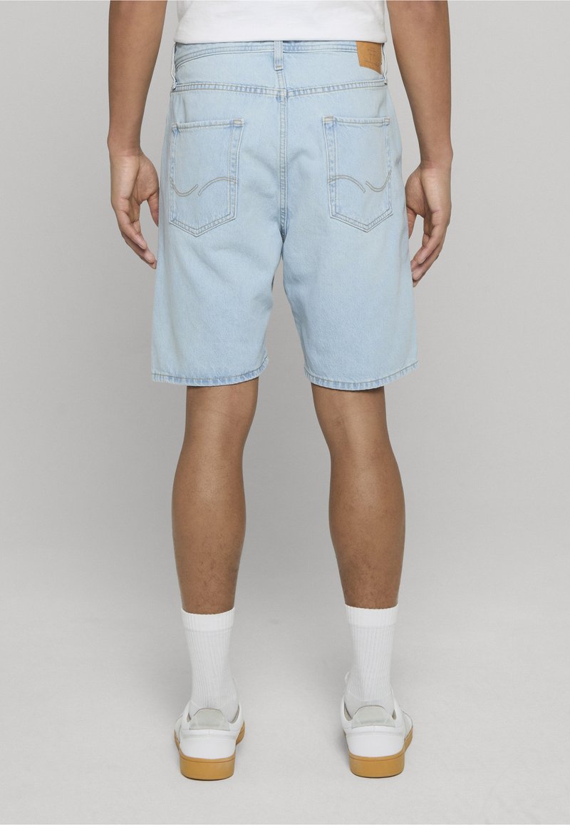 Mann trägt hellblaue Jeansshorts, weiße Socken und weiße Turnschuhe, steht mit locker an den Seiten hängenden Händen vor einem schlichten Hintergrund.