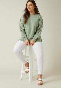 Sudadera verde claro con diseños cortados intrincados en las mangas, combinada con pantalones blancos y sandalias blancas, sentada en un taburete.