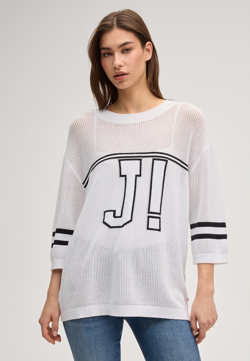 Jeune femme portant un pull en maille blanche ample avec des rayures noires et un lettrage audacieux « J! », associée à un jean bleu, fond neutre.