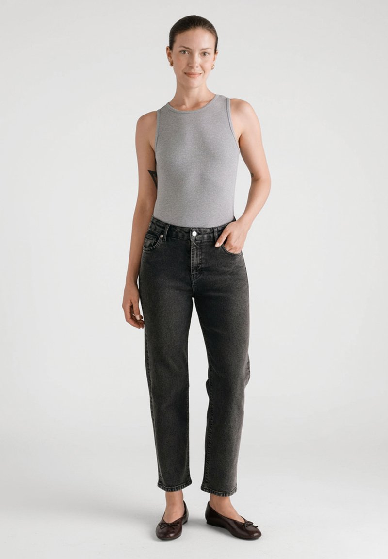 Szary bezrękawnik typu body, czarne jeansy z wysokim stanem i ciemnobrązowe baleriny, stojąca na neutralnym tle.