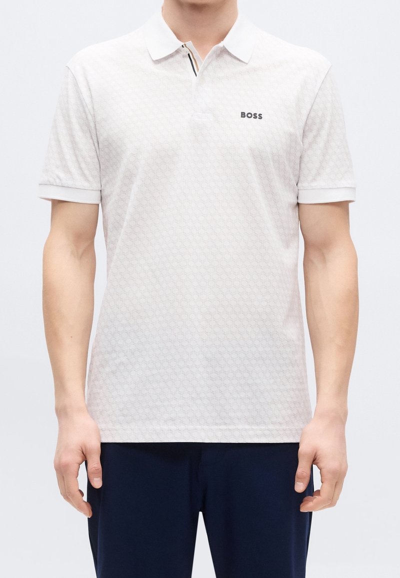 Homme portant un polo blanc à motifs avec le logo "BOSS" sur la poitrine et un pantalon foncé, debout, bras détendus.
