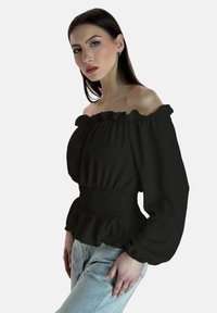 Blusa nera scollata sulle spalle con scollatura con volant, vita arricciata e maniche lunghe a sbuffo, abbinata a jeans in denim blu chiaro.