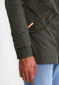 Primer plano de una mano junto a la solapa del bolsillo de una chaqueta Superdry color verde oliva, combinada con unos vaqueros azul claro.