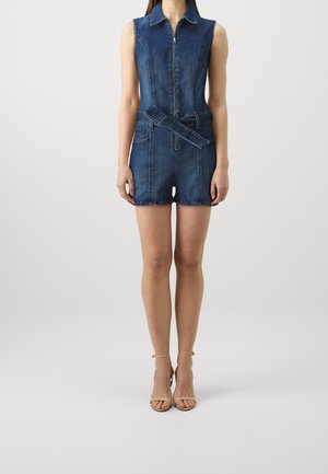 Denim ærmeløs dragt med lynlås foran, bælte i taljen, to sidelommer og lys blå vask, stylet med beige sandaler med stropper.
