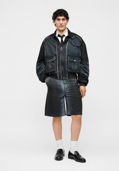 Άνδρας που φοράει μαύρο και σκούρο μπλε μπουφάν με φερμουάρ και μεγάλες τσέπες, σκούρα μπλε βερμούδα μέχρι το γόνατο, λευκές κάλτσες και μαύρα επίσημα παπούτσια.