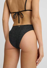 Slip inferiore di bikini nero con finitura texturizzata, forma alta e dettaglio in metallo dorato in vita. Presenta laccetti regolabili sul retro.